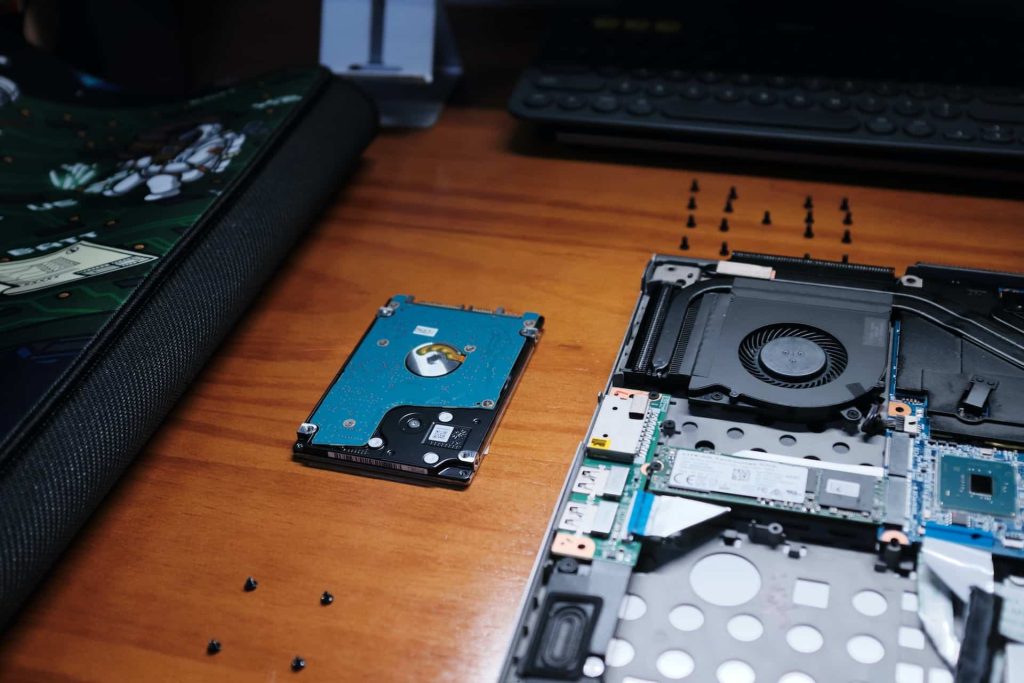 A computer hard drive and cooling fan lie on a wooden desk. The laptop is open, exposing its internal components, with several small screws scattered around. A keyboard is visible in the background, ready for its next step: electronic recycling at the drop-off box in Atlanta, GA.