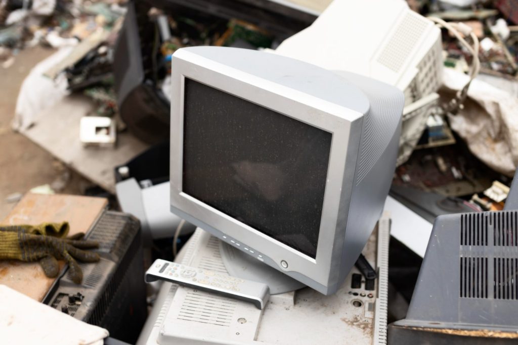 A pile of electronic waste lies discarded outdoors in Atlanta, GA, featuring a gray CRT monitor, a remote control, and various dismantled computer parts. On the left, a pair of gloves signals the need for responsible recycling to protect the environment.