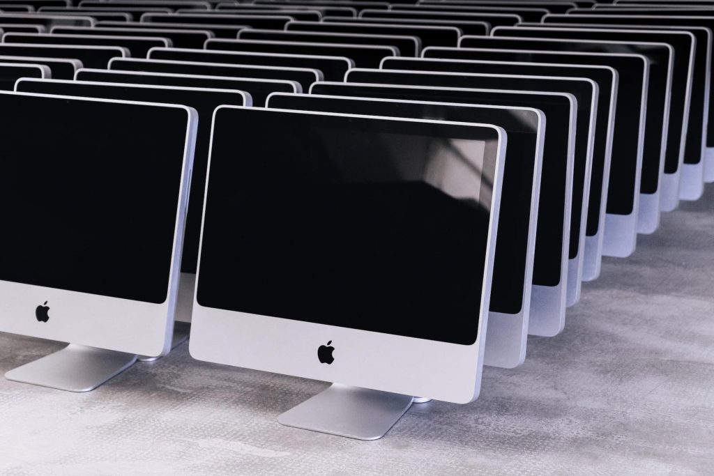 A large number of silver desktop computers with black screens are neatly arranged in rows on a gray floor, embodying modernity. Each has a thin design with a logo on the front, highlighting Atlanta's commitment to electronics recycling and waste reduction initiatives.