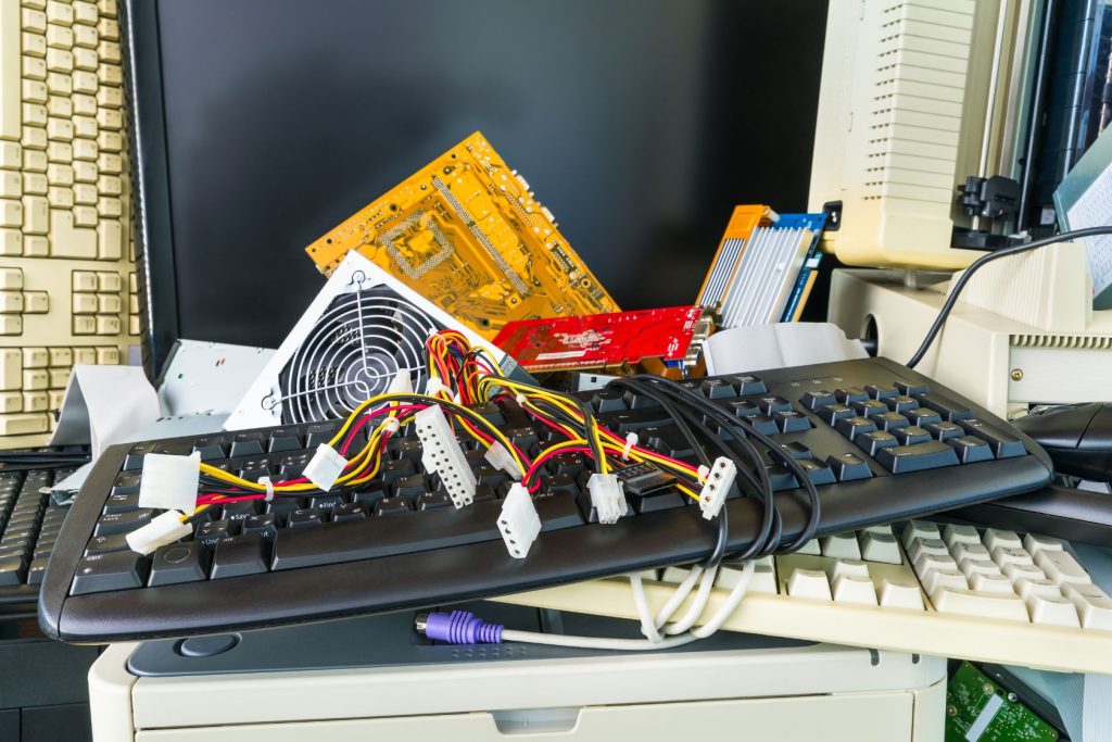 A cluttered pile of old computer components, including keyboards, a cooling fan, and a graphics card, stacked in front of a black monitor—a scene typical for those involved in electronic recycling in Atlanta, GA.