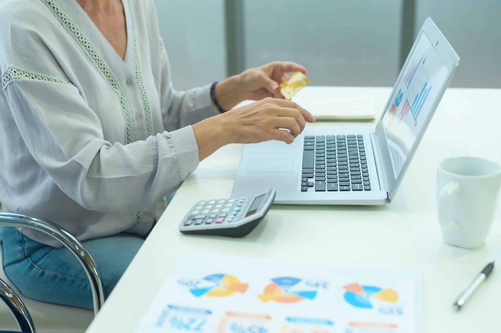 A person working in a modern Atlanta, GA office uses a credit card on a laptop at their desk. Nearby, a calculator, mug, and paper with colorful charts capture attention—a glimpse into efficiently blending technology with work.