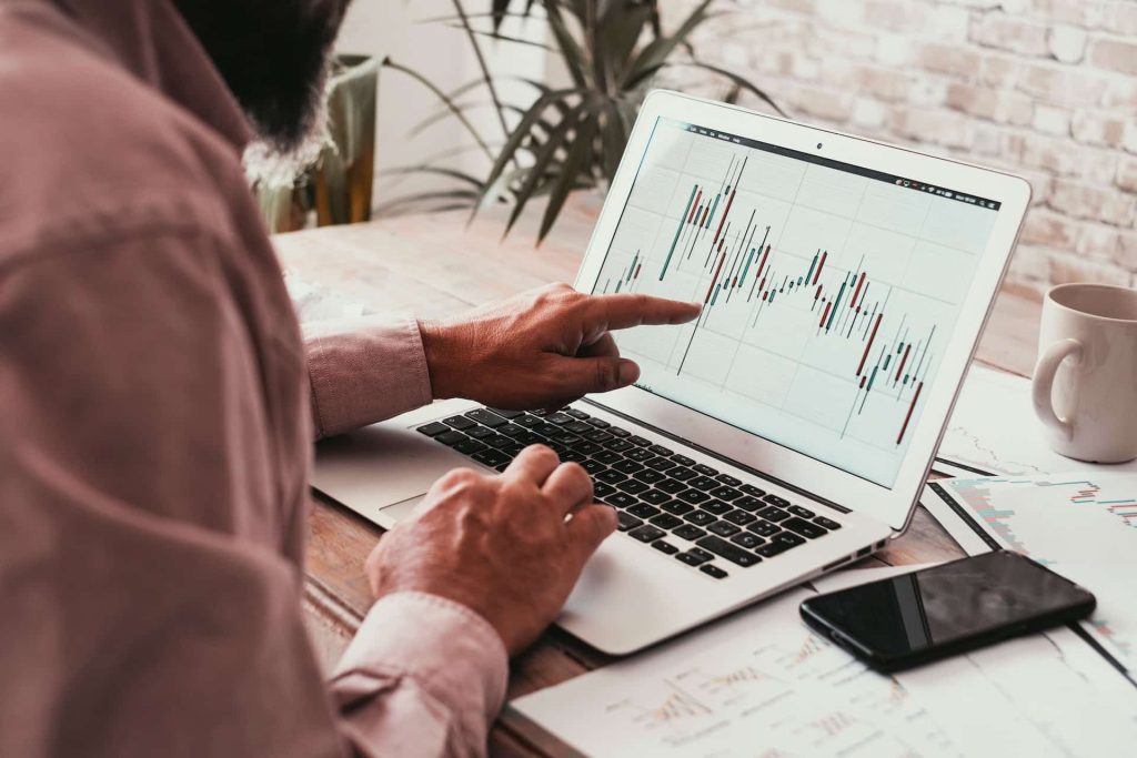 A person analyzing data on a laptop, with a financial chart displayed prominently. The bustling workspace, reminiscent of Electronics Atlanta GA, features scattered papers and a smartphone on a wooden desk. A plant adds a touch of greenery in the background.