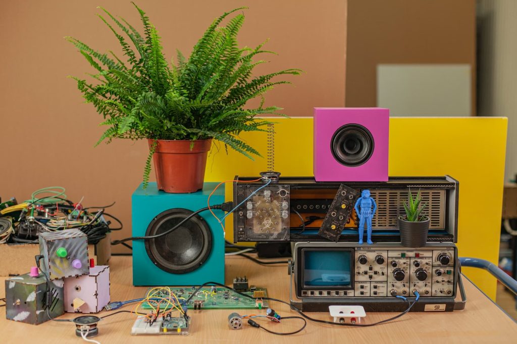 A colorful, eclectic arrangement featuring potted ferns, vintage electronics sourced from Electronic Waste in Atlanta GA, a speaker in a pink box, a small blue figurine, and various circuit boards with wires—all on a wooden surface against a brown and yellow backdrop.