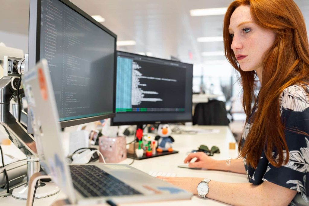 A woman with long red hair is deeply focused at her desk in a bright office environment filled with gadgets and electronics, perhaps sourced from Atlanta, GA. Multiple monitors display complex code while a toy penguin keeps her company amidst the digital hubbub.