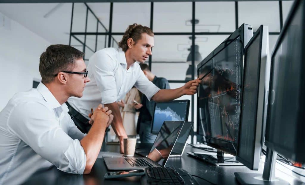 In a modern Atlanta office, two men analyze data on multiple computer screens. One, standing, points at a monitor while the other observes. Both are focused, surrounded by laptops and team members—it's a hub for electronics innovation with eco-friendly drop-off boxes nearby.