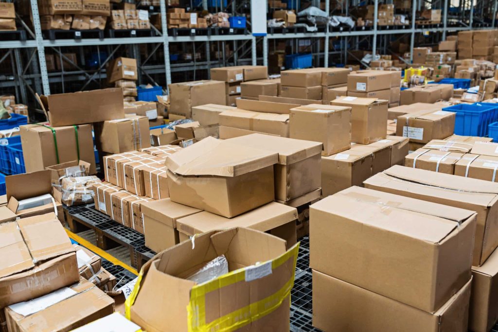 A warehouse in Atlanta, GA is filled with numerous brown cardboard boxes of various sizes stacked on shelves and scattered on the floor. Blue plastic containers and electronic waste drop-off boxes are also visible in the background among the organized shelves.