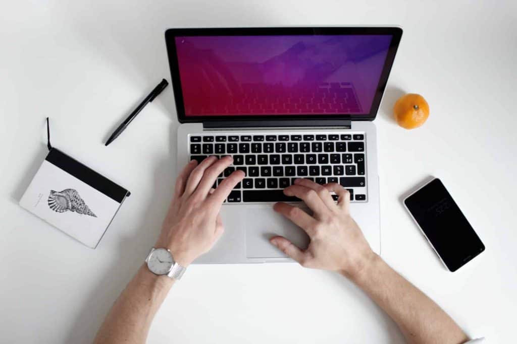 A person types on a laptop with a blank screen, surrounded by a closed shell-design notebook, a pen, and a smartphone. An orange rests nearby on the white desk in Atlanta, GA, where electronic recycling is thoughtfully supported with convenient drop-off boxes. Hands are visible, one adorned with a watch.