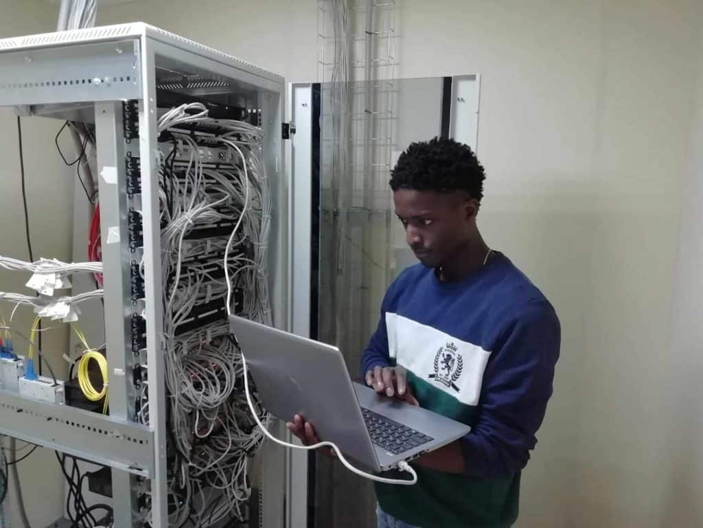 A person stands in front of an open server rack filled with cables, focused on working on a laptop. The setting appears to be a server room or data center, underscoring the importance of proper electronics management like electronic recycling in Atlanta, GA.