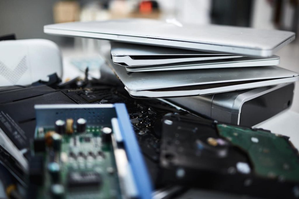 A close-up of several stacked laptops, a partially disassembled hard drive, and various electronic components scattered on a table in Atlanta GA. The image conveys a sense of repair or recycling of electronic waste.