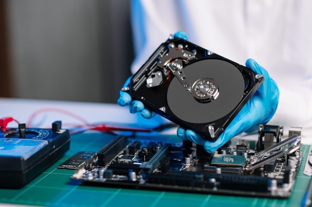 A person wearing blue gloves holds a hard drive over a computer motherboard on a green mat, surrounded by a multimeter and tools, indicating electronic repair. The scene is reminiscent of an Electronics Atlanta GA workshop where precision and tech care are paramount.