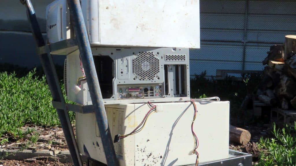 A pile of old computer towers and equipment, ready for drop-off, is stacked outdoors next to a ladder. The exposed circuit boards and wires gleam in the sunny Atlanta, GA garden, with logs casting shadows in the background.