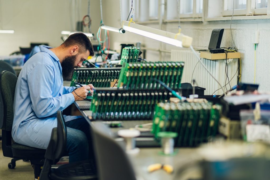 A technician in a blue lab coat is meticulously assembling circuit boards at a well-organized workstation in Atlanta, GA. Surrounded by various electronic components and equipment, the scene highlights the city's pivotal role in electronics innovation.