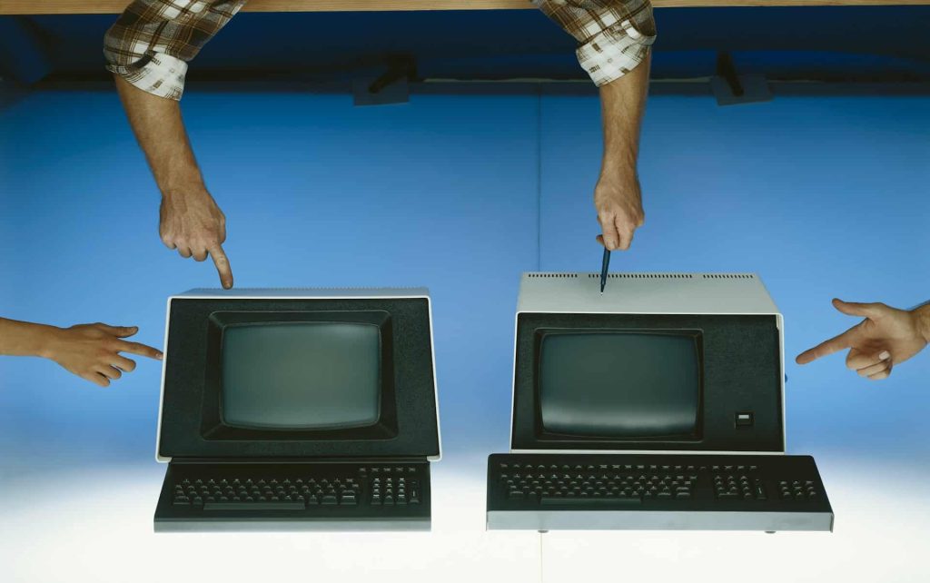 Two vintage computers are positioned side by side, with hands pointing at each one, against a blue background. Each monitor has a keyboard in front. These relics remind us of the growing pile of electronic waste in Atlanta, GA. Consider using a local drop-off box for proper disposal.