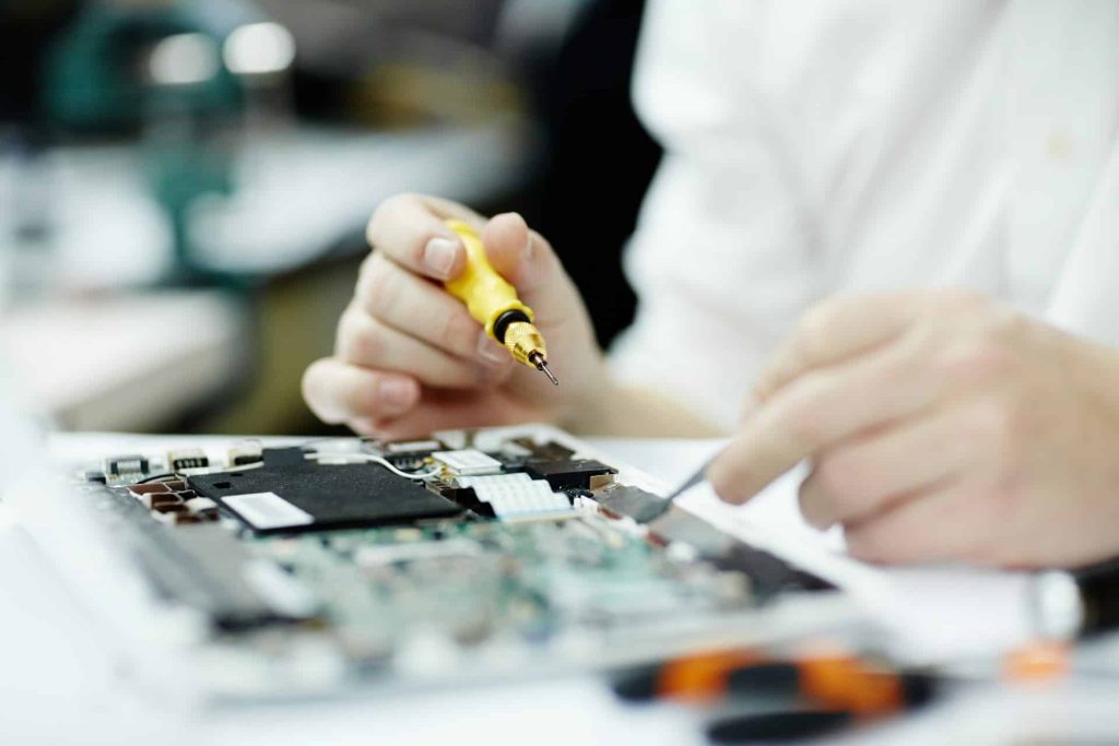 A person is carefully using a soldering iron on a circuit board, their hands deftly holding the tool and a small component. The blurred background emphasizes the precision work on the electronic device, much like meticulous electronic recycling in Atlanta GA aims to conserve resources.