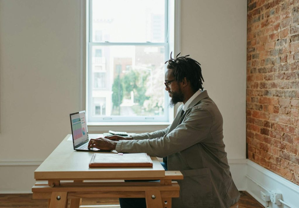 A man sits at a wooden desk, working on a laptop in a room with a large window and exposed brick wall, resembling an electronics workspace in Atlanta GA. The scene suggests a relaxed, focused environment, perhaps near an Electronic Recycling drop-off box.