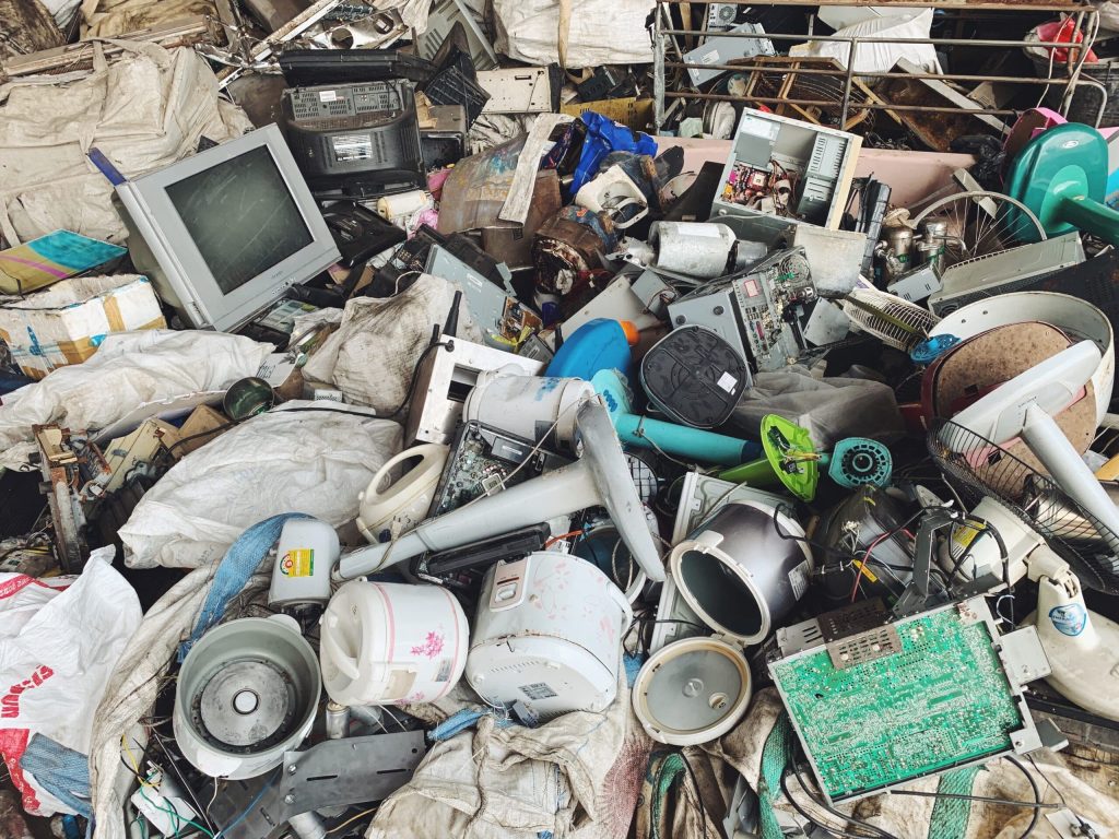 A large pile of electronic waste, including old monitors, circuit boards, speakers, and various household gadgets, is heaped together. The background reveals more discarded items and fabric bags awaiting their fate at an Electronic Recycling facility in Atlanta GA.