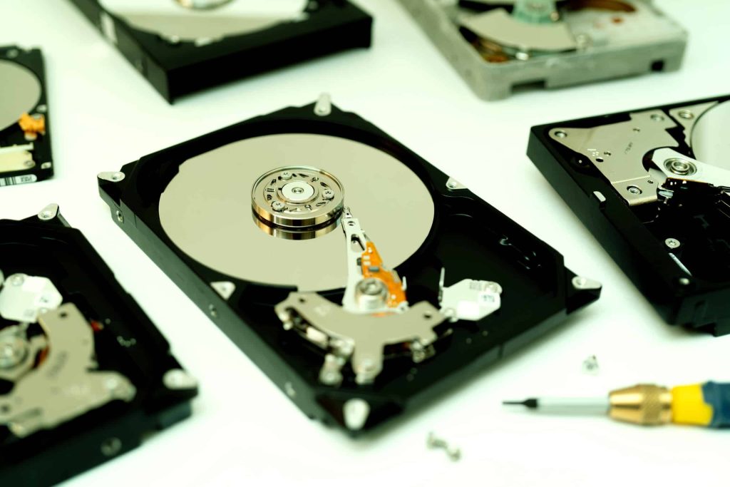 Several open hard drives are arranged on a white surface, with visible platters and read/write heads. A screwdriver and small screws are scattered around, indicating disassembly, reminiscent of an electronics workshop in Atlanta GA.