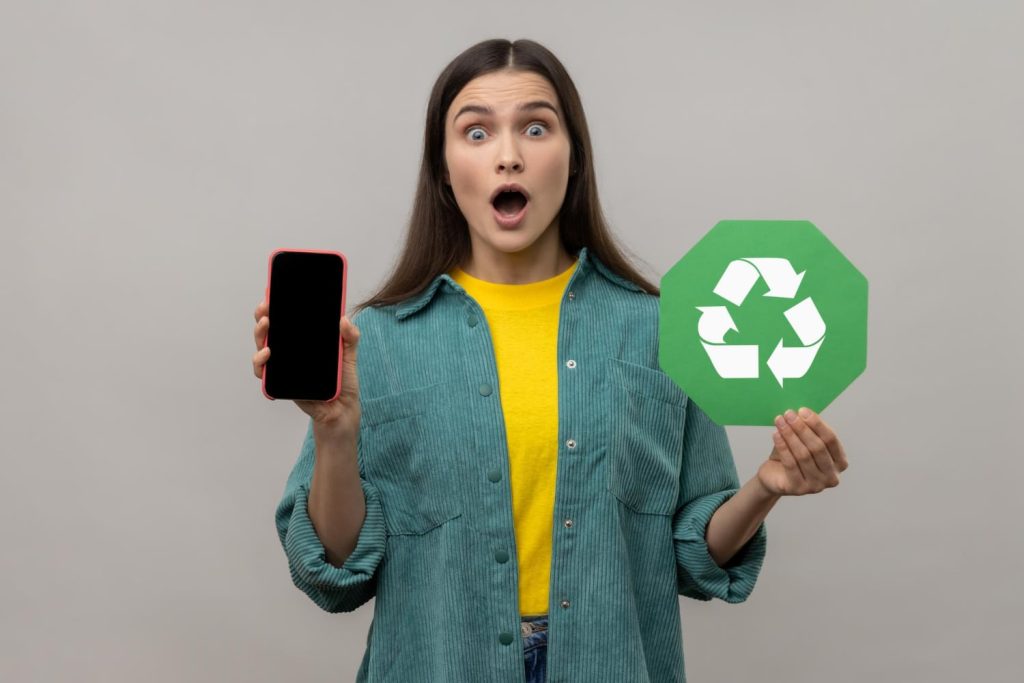 A surprised woman holds a smartphone in one hand and a green recycling symbol in the other, highlighting electronic recycling in Atlanta, GA. She wears a green shirt over a yellow top, standing against a plain background.