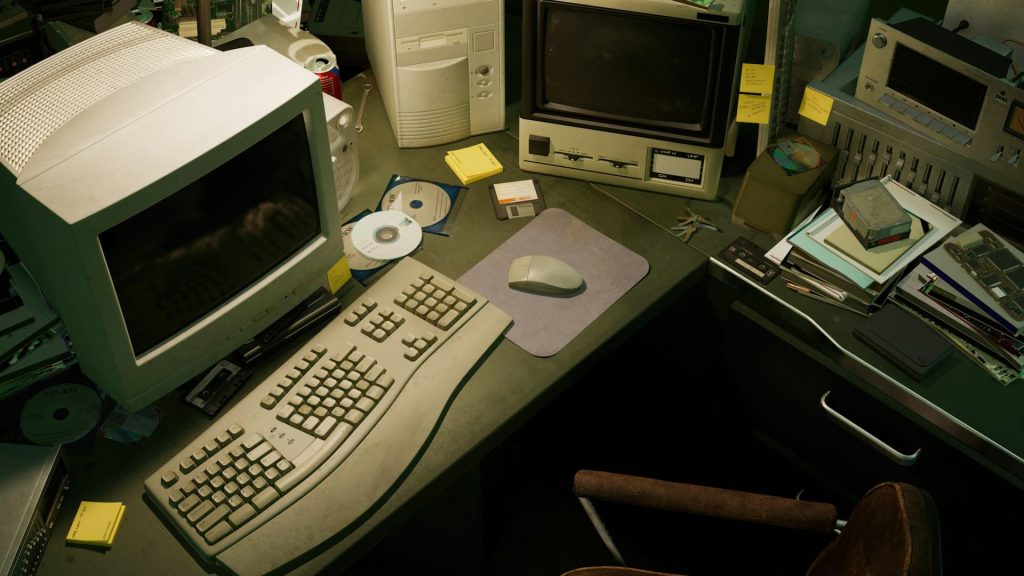 A cluttered desk with vintage computer equipment, including a CRT monitor, keyboard, mouse, and floppy disks. Surrounding items like CDs and sticky notes form a retro-tech atmosphere reminiscent of the bygone era of electronics in Atlanta GA.