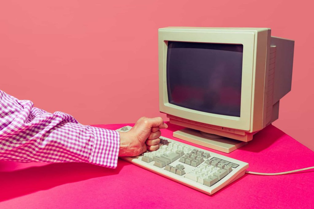 A person in a checkered shirt fist-bumps an old-style beige computer keyboard and monitor against a vibrant pink background, embodying the spirit of electronic recycling in Atlanta, GA.