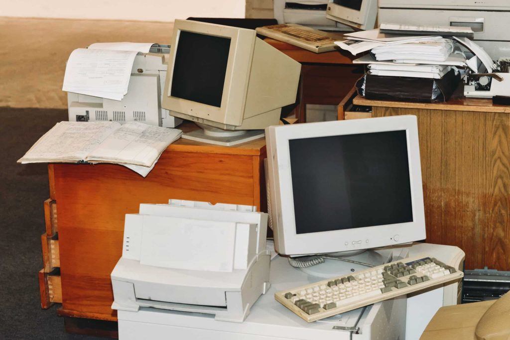 A cluttered office desk with two vintage CRT monitors, a keyboard, a printer, and stacks of papers evokes a retro tech workspace vibe. Ideal for those into electronics in Atlanta GA, this setup on wooden furniture takes you back to when electronic recycling was unheard of.