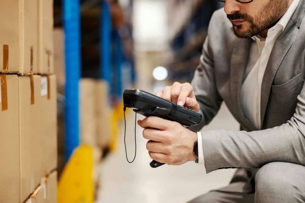 A man in a suit crouches in a warehouse, using a handheld scanner beside a row of stacked drop-off boxes for electronic recycling. The background is filled with industrial shelves and more boxes, illustrating the bustling environment of electronics management in Atlanta, GA.