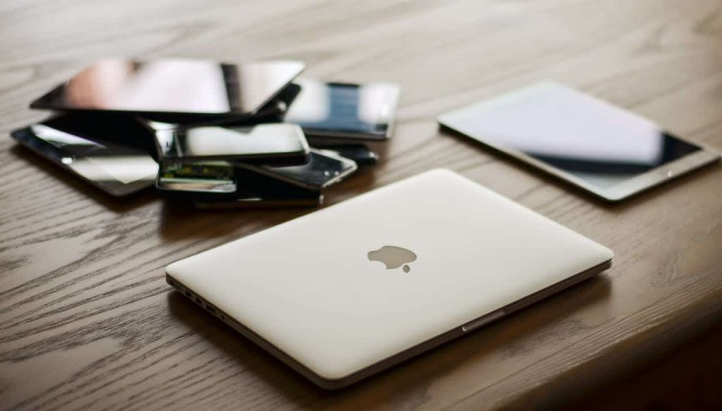 A sleek silver laptop with an apple logo on its closed lid sits on a wooden table. Behind it is a stack of various smartphones and tablets, all with black screens, ready for electronic recycling in Atlanta, GA.