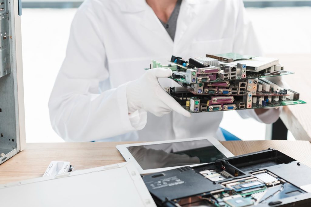 A person in a white lab coat and gloves holds several stackable computer circuit boards, poised for electronic recycling in Atlanta, GA. Nearby, electronic devices and disassembled parts are scattered on a wooden table in the bright workspace.