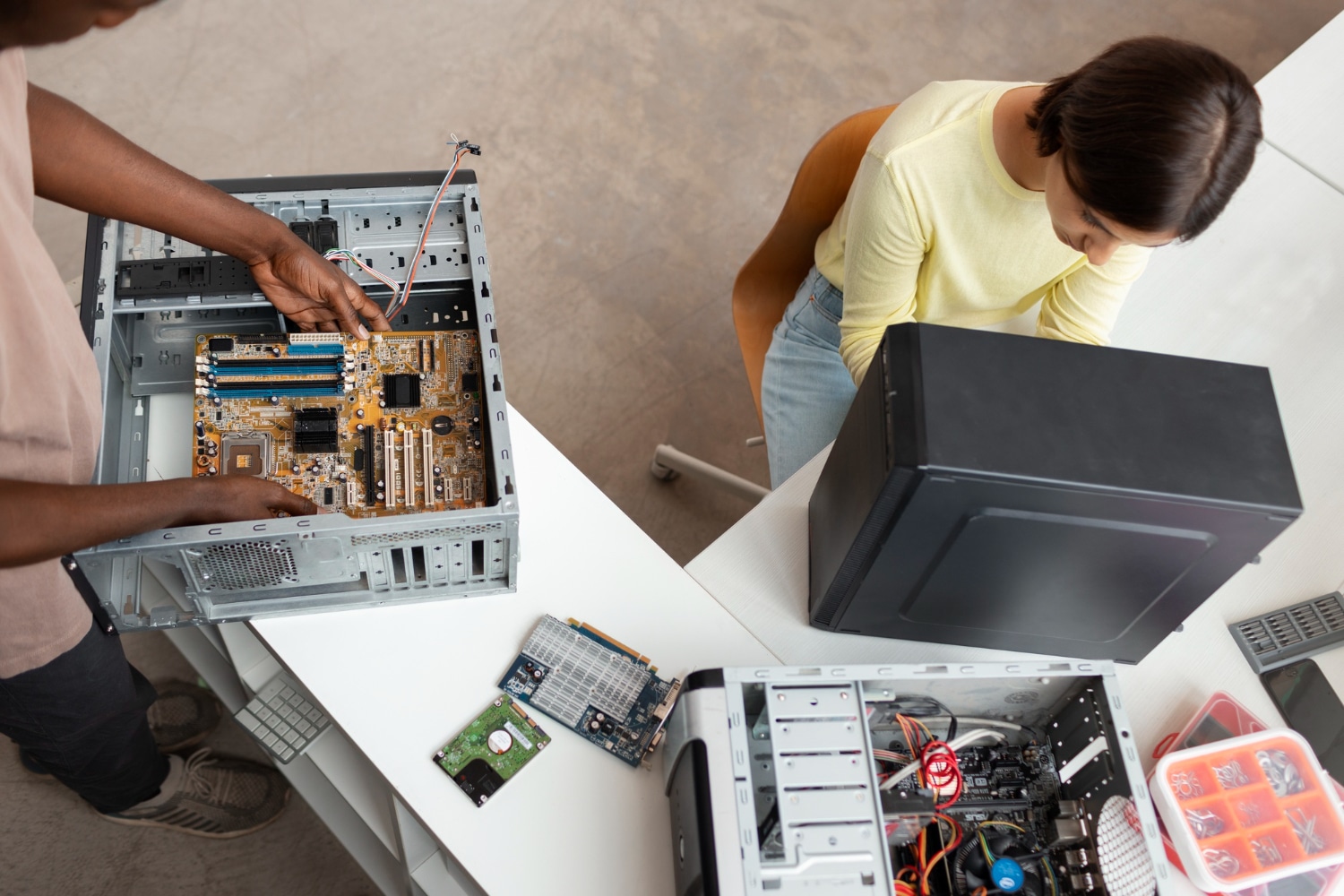 A person is assembling a computer, installing components on a motherboard inside a desktop case. Nearby, someone examines a computer case labeled "Electronic Recycling Atlanta GA." Various computer parts and tools are scattered on the table.