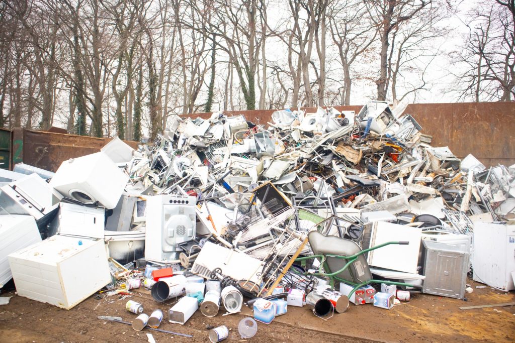 A large pile of discarded household appliances, including washing machines, ovens, and microwaves, is stacked in an outdoor junkyard surrounded by bare trees. Various metal and plastic debris lies scattered around at the Electronic Recycling Atlanta GA site.