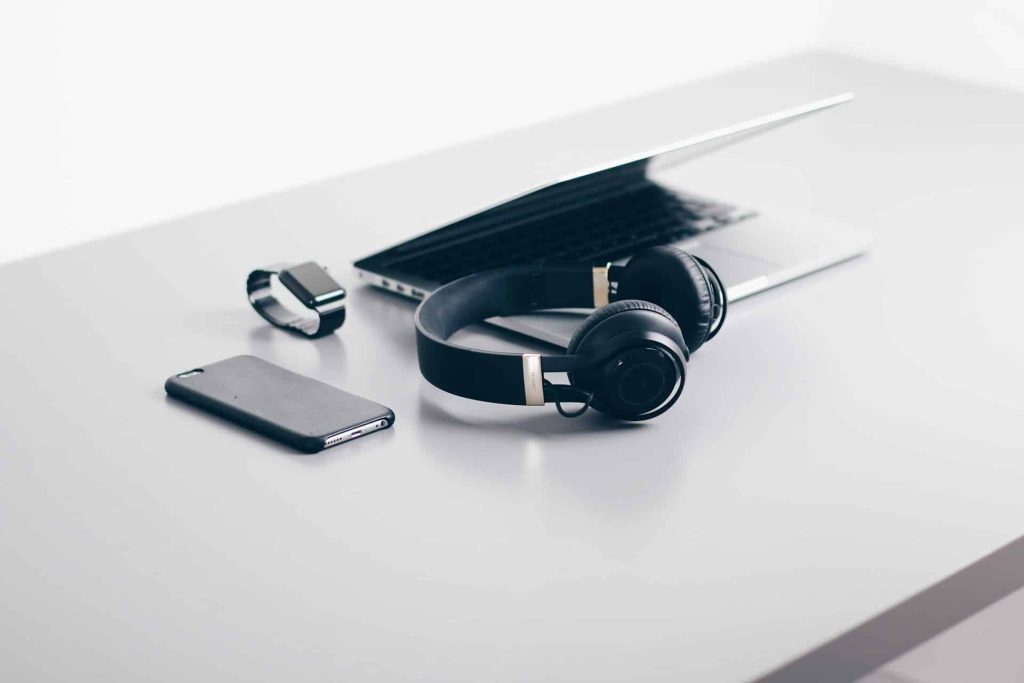 A minimalist desk setup in Atlanta, GA, showcases a laptop partially closed, a pair of black wireless headphones, a smartphone, and a smartwatch—all placed neatly on a smooth, light gray surface. Consider using an electronics drop-off box for recycling and sustainability in the Atlanta area.