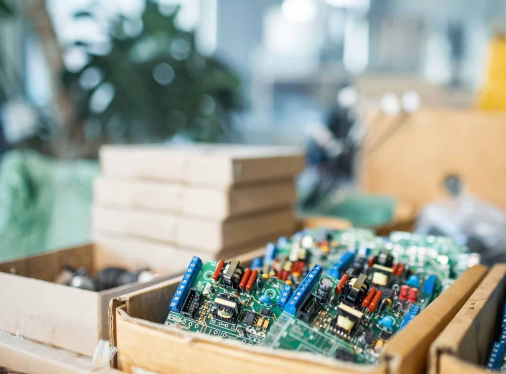 Close-up of electronic circuit boards inside a cardboard box on a cluttered workbench. The scene evokes the busy workshop vibes of Electronics Atlanta GA, with a blurred background featuring stacked boxes and a plant.