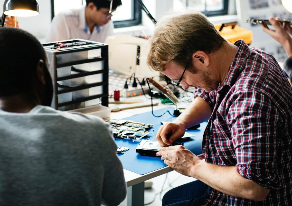 A group of people is working intently on various electronic devices at a table in Atlanta, GA. One man in the foreground focuses on assembling or repairing a gadget. The background shows others engaged in similar activities with tools and components scattered around, part of an electronics recycling effort.