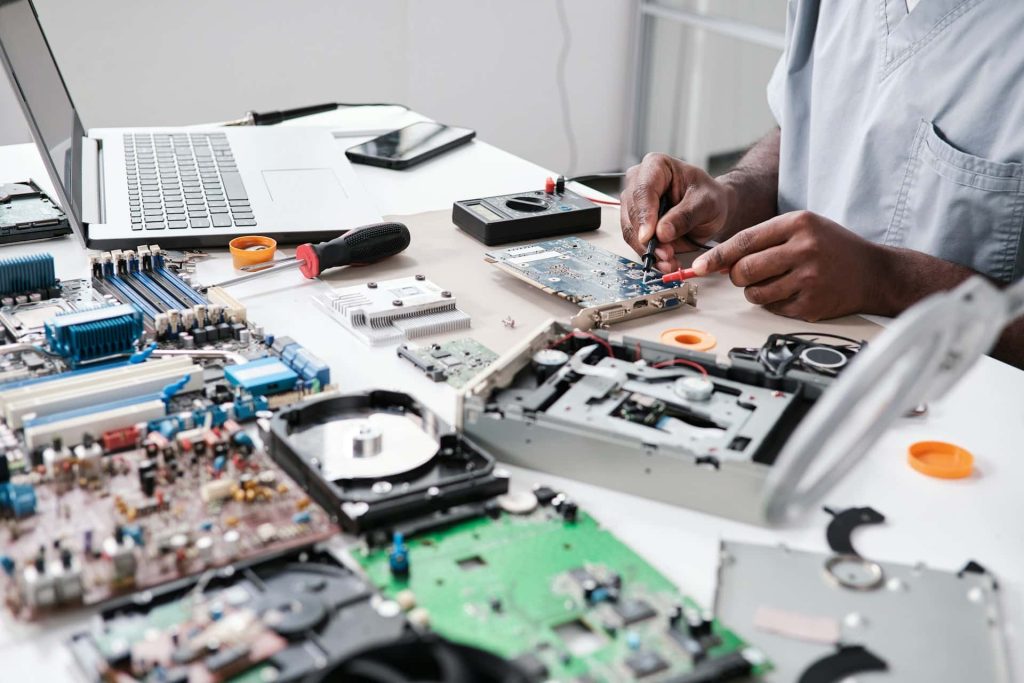 A person is working on electronic components at a cluttered desk, filled with circuit boards, a laptop, tools, and various hardware parts. They focus intently on a circuit board, perhaps considering the potential for recycling or disposal through Electronic Waste Atlanta GA.