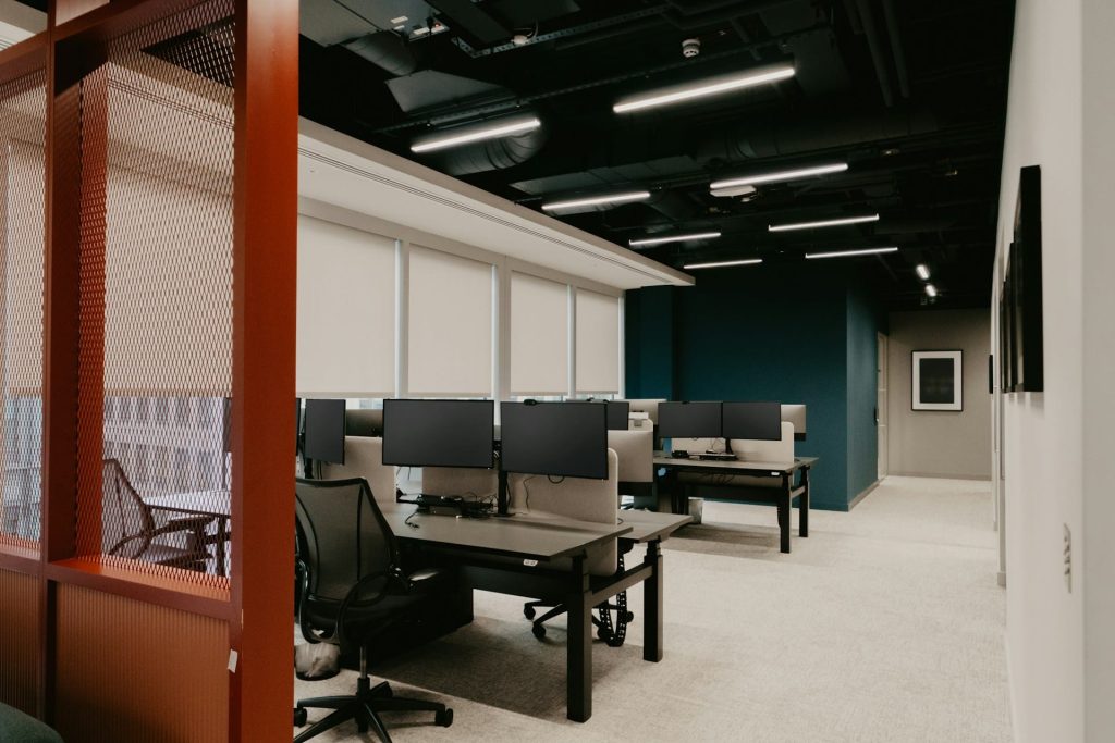 A modern, empty office space with multiple workstations awaits in the heart of Electronics Atlanta GA. Each workstation has a desk, an office chair, and dual monitors. The room is well-lit with ceiling lights, and the windows have roller blinds partially drawn. A partition with a mesh panel is visible.