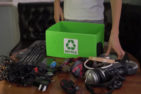 A person is carefully sorting a collection of electronic waste for e-waste disposal into a green recycling box on a table. The surface holds various items like a keyboard, headphones, cords, and small gadgets ready for eco-friendly recycling.