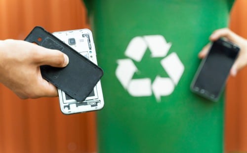 A person wearing a green shirt with a white recycling symbol holds a disassembled smartphone and another phone. The background is blurred, emphasizing electronics recycling and the importance of responsible electronics waste disposal through the prominent recycling symbol.