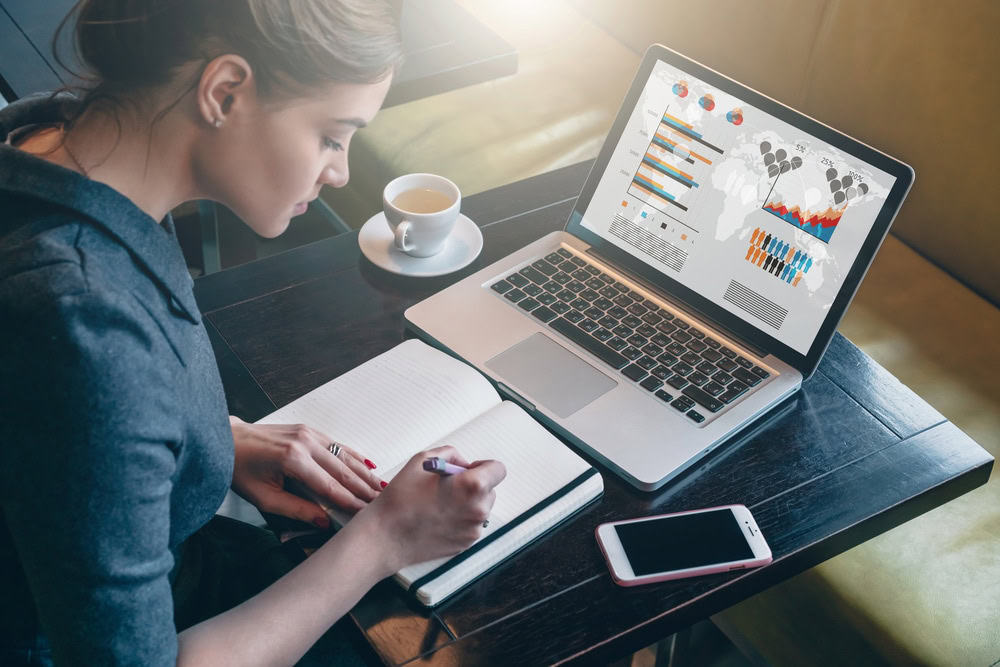 A woman sits at a table, writing in a notebook while looking at a laptop displaying colorful graphs and charts about electronics recycling. A cup of coffee and a smartphone are on the table. Bright sunlight filters in, creating a focused atmosphere.