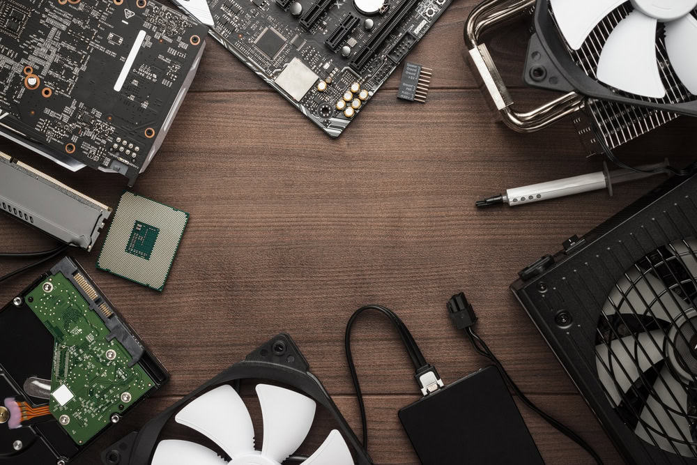 Computer components arranged around a wooden surface, featuring motherboards, fans, a processor, a hard drive, and a small black external device. A screwdriver and thermal paste are also visible. The setup highlights the importance of electronics recycling to reduce waste and repurpose parts.