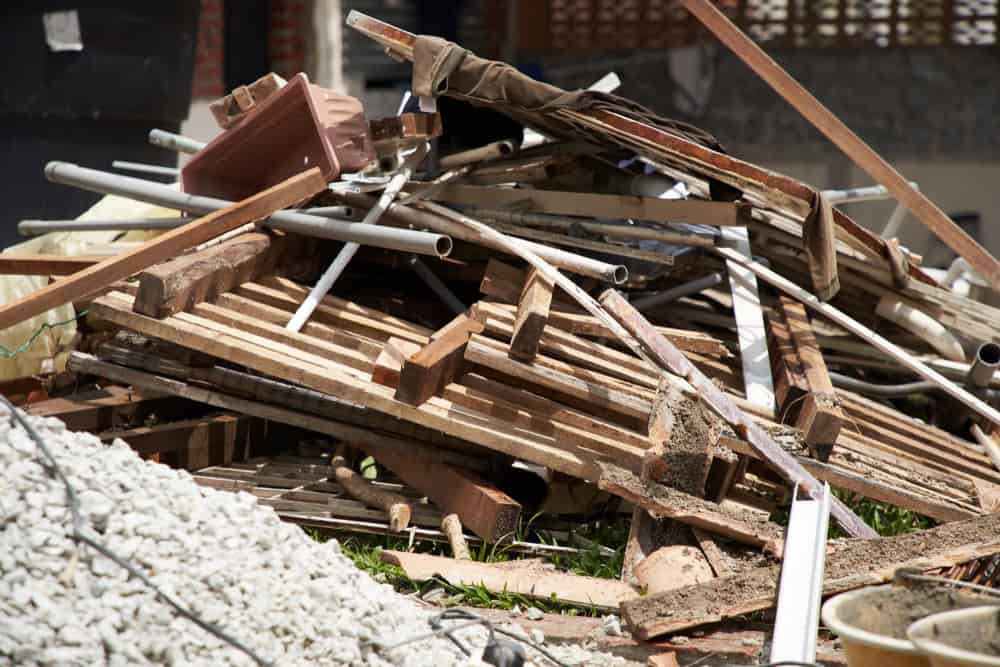 A pile of construction debris, including wooden planks, pipes, bricks, and other materials, is seen on the ground. There are some weeds growing around the pile, and a building is visible in the background.