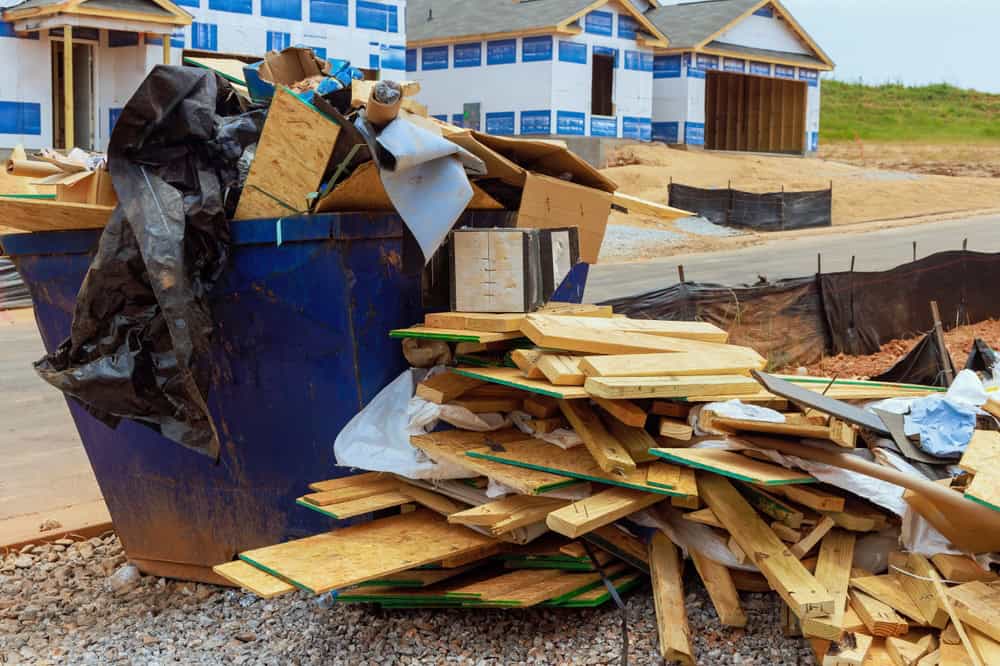 A blue dumpster overflowing with construction debris, including wooden planks and black plastic, is on the gravel road of a building site. In the background, partially constructed houses are visible with protective coverings.