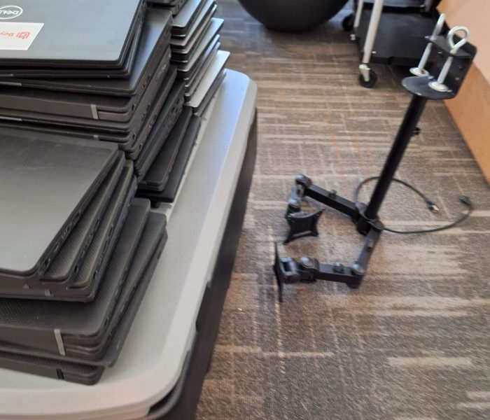 A stack of laptops rests on a portable cart, awaiting electronics recycling. Nearby, a deflated exercise ball and a tripod stand are visible on the carpeted floor, surrounded by various office and exercise equipment.