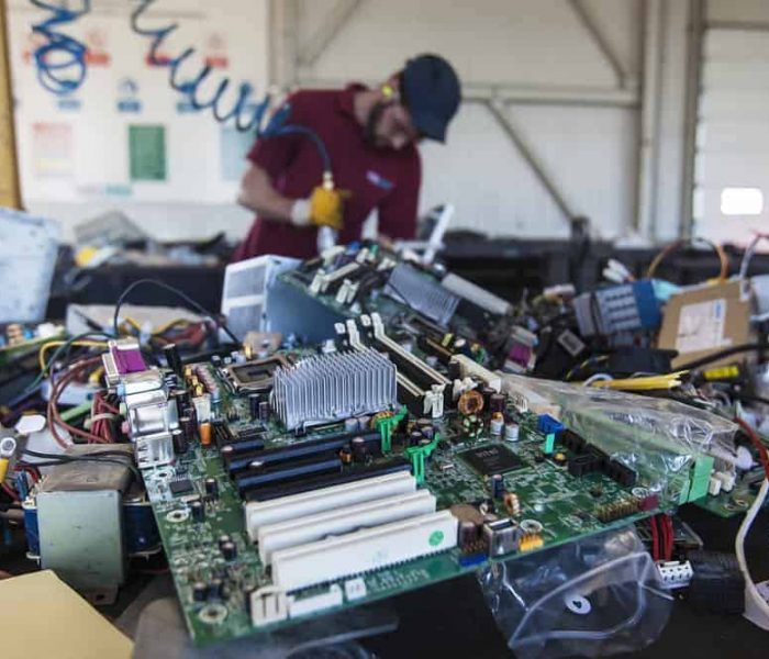 Close-up of a pile of electronic waste, featuring a motherboard in focus. In the background, a person wearing gloves and a hat is working with tools in an industrial setting, slightly blurred—a scene highlighting the importance of efficient E-waste disposal.