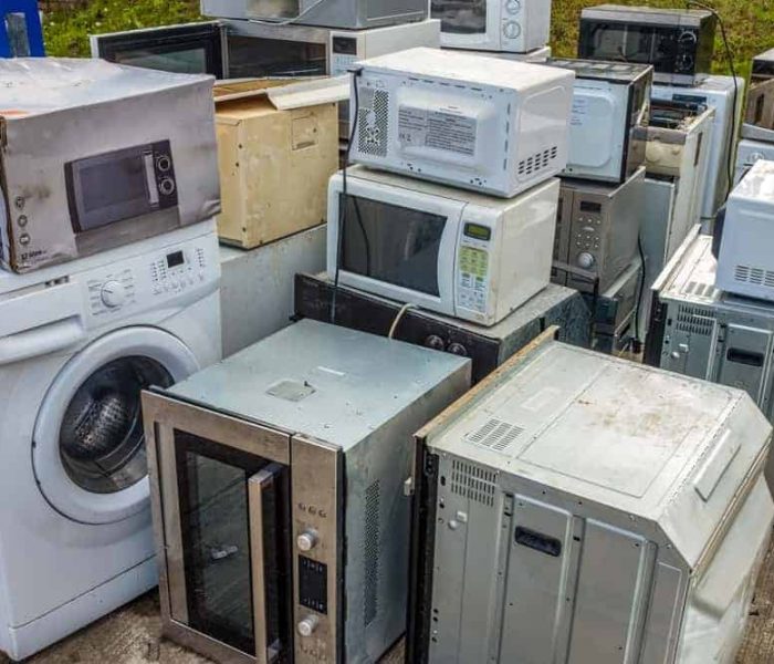 A pile of discarded home appliances, including microwave ovens, a washing machine, and other electronic devices, is stacked outdoors. Weathered and in disarray, these items highlight the importance of electronics waste disposal as they await recycling or disposal.