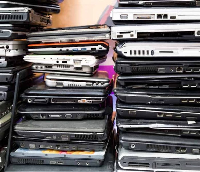 Stacks of various old laptops piled closely together, showcasing a diverse range of models and sizes. The laptops are stacked in two columns, highlighting the importance of electronics recycling and the variety of ports and designs on each device.